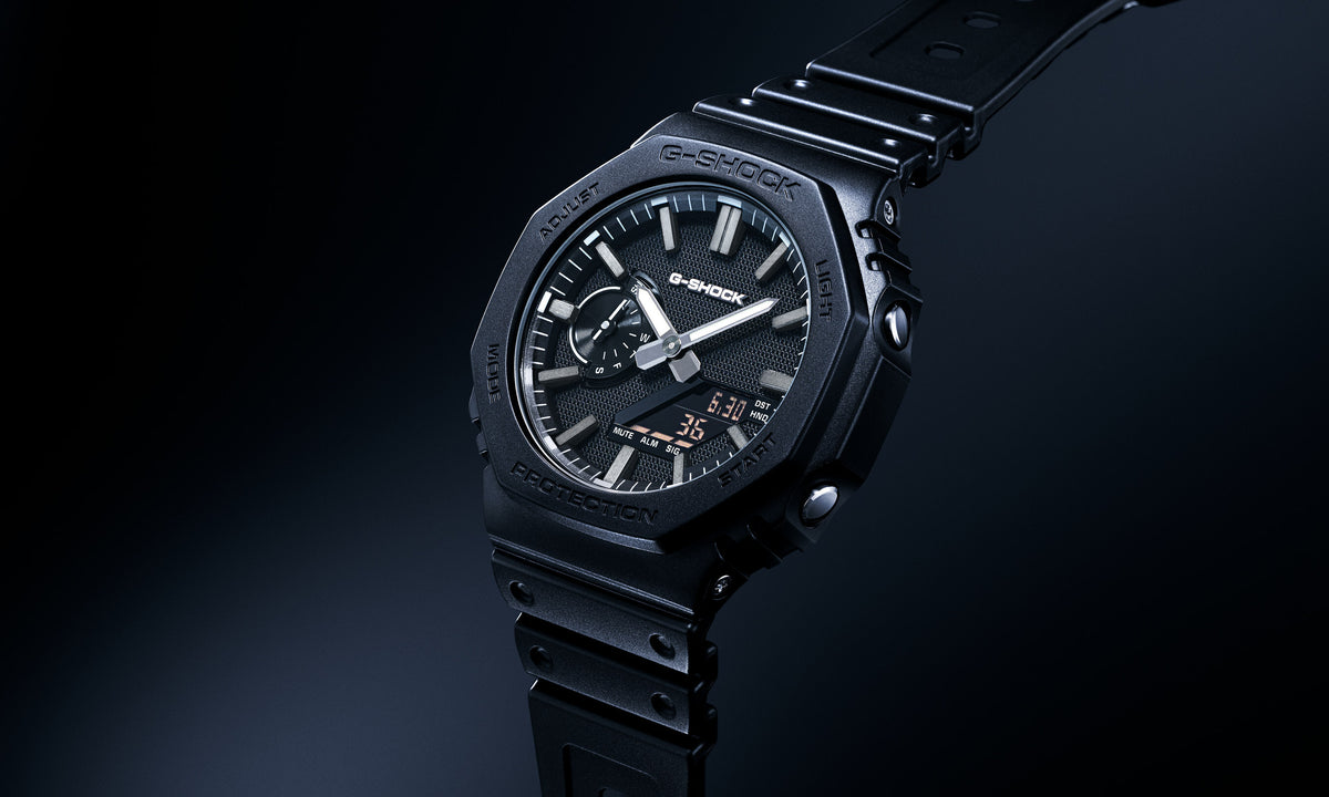 A black Casio G-Shock watch with a dark dial, silver-tone hands, and orange hour markers, set against a dark studio background.