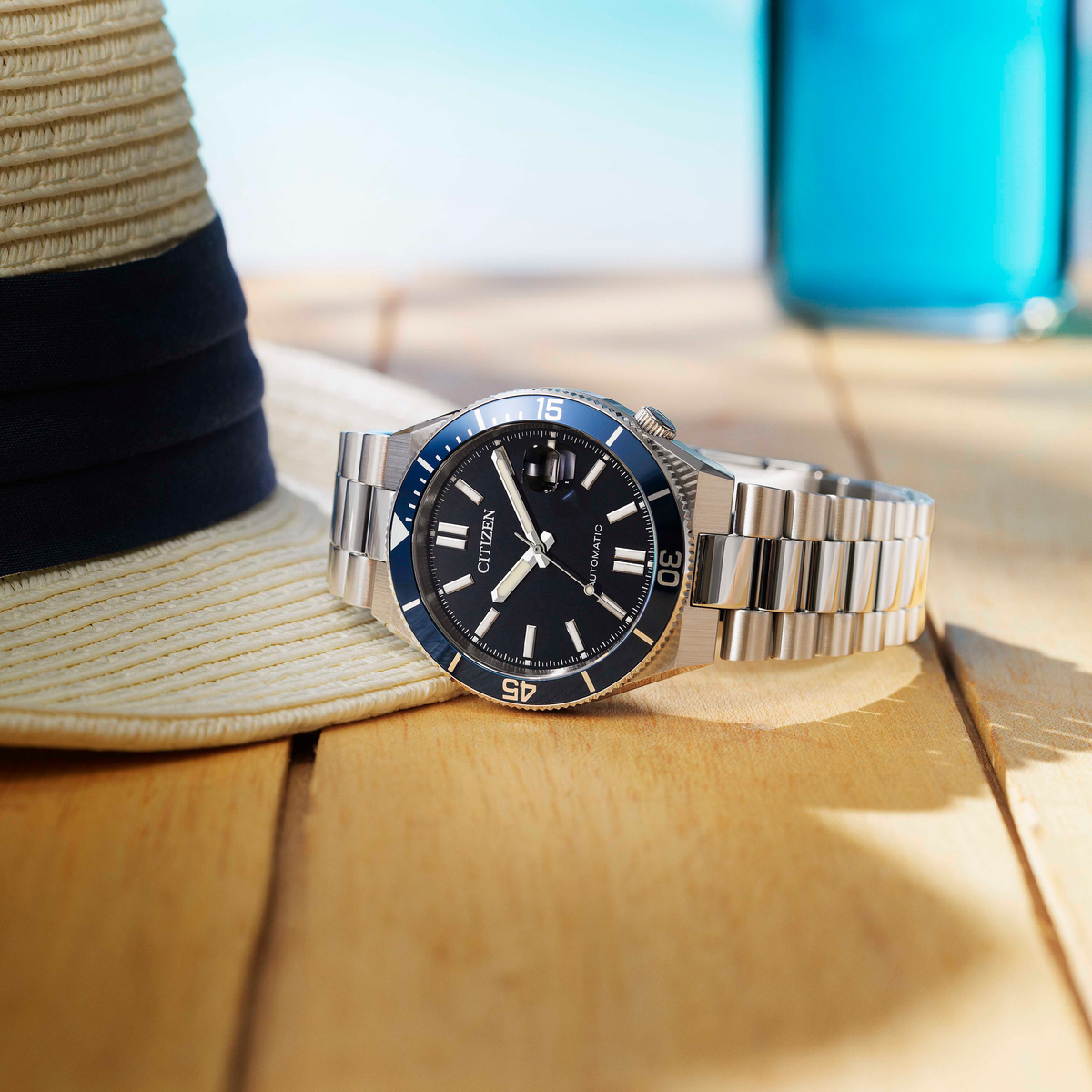 A Citizen stainless steel watch with a blue bezel and black dial, resting on a wooden surface next to a straw hat and a blue cup.